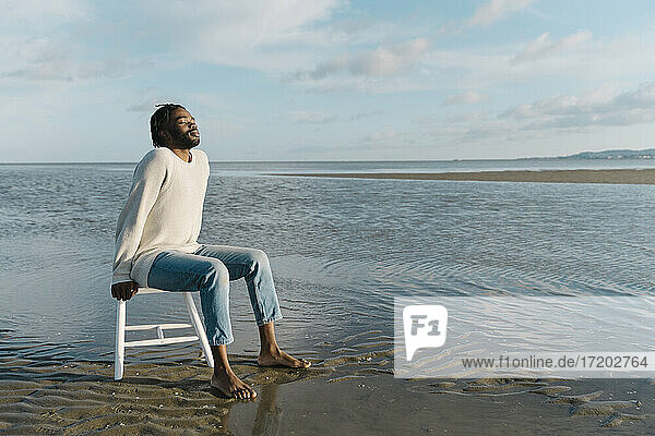 Nachdenklicher junger Mann mit geschlossenen Augen sitzt auf einem weißen Hocker am Strand bei Sonnenuntergang