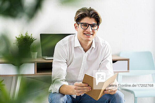 Lächelnder junger Mann mit Buch im Wohnzimmer sitzend