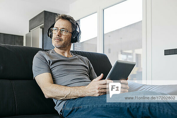 Älterer Mann mit digitalem Tablet  der wegschaut  während er auf dem Sofa im Wohnzimmer Musik über Kopfhörer hört