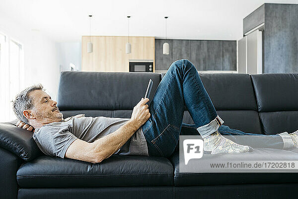 Älterer Mann schaut auf ein digitales Tablet  während er auf dem Sofa im Wohnzimmer liegt
