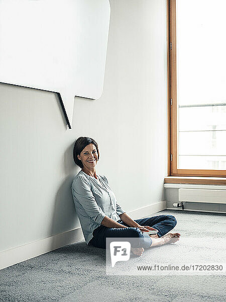 Lächelnde Unternehmerin sitzt auf dem Boden in einem Büro an der Wand