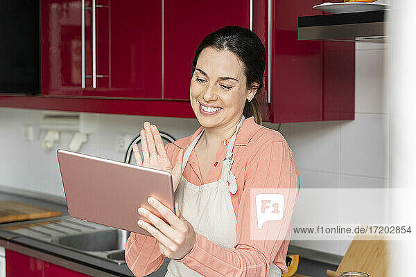 Schöne Frau winkt mit der Hand während eines Videoanrufs auf einem digitalen Tablet in der Küche zu Hause