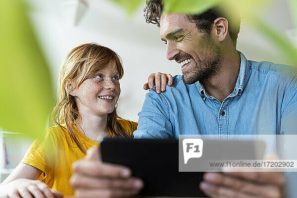 Vater und süße rothaarige Tochter lächeln sich gegenseitig an  während sie ein digitales Tablet im Wohnzimmer halten