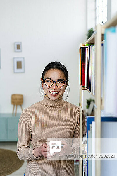 Junge lächelnde Frau mit Mobiltelefon  die zu Hause am Bücherregal steht