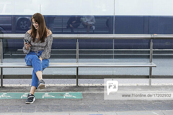 Lächelnde junge Frau  die ein Smartphone benutzt  während sie auf einer Bank an einer Straßenbahnhaltestelle sitzt