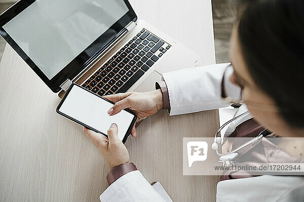 Female doctor using mobile phone by laptop at desk