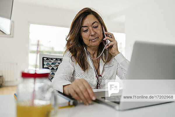 Unternehmerin  die mit ihrem Smartphone telefoniert  während sie zu Hause mit ihrem Laptop auf dem Schreibtisch sitzt