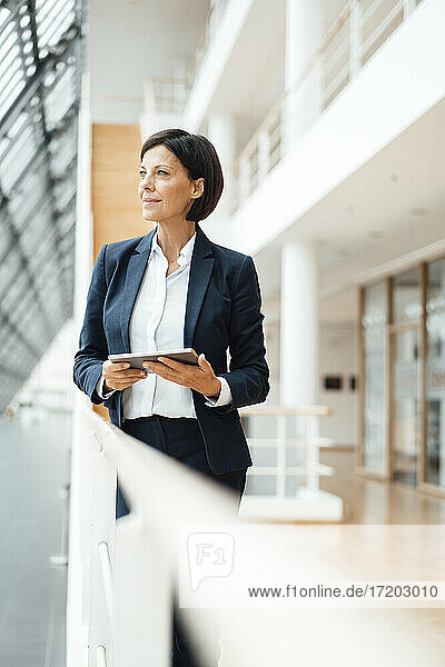 Nachdenkliche Geschäftsfrau mit digitalem Tablet  die am Geländer im Korridor wegschaut