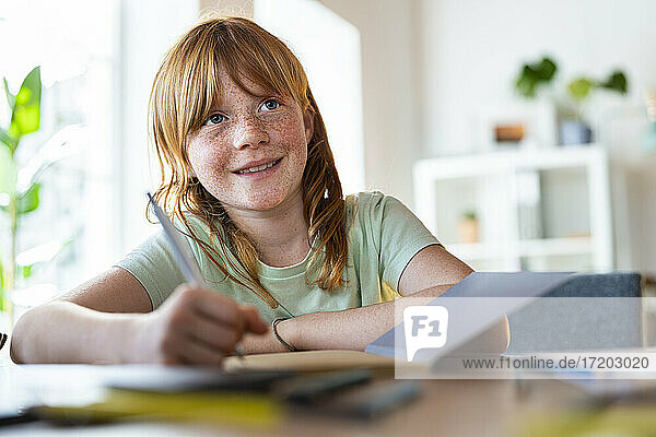 Cute smiling redhead girl looking away while doing homework at home
