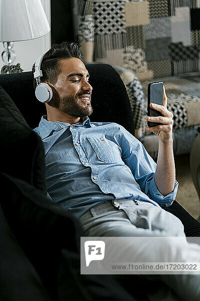 Smiling man wearing headphones using mobile phone while lying on sofa at home