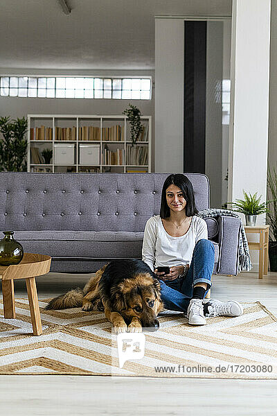 Smiling woman sitting with dog on carpet against sofa in living room