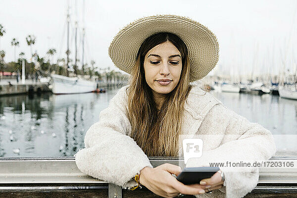 Junge Frau mit Hut benutzt ein Mobiltelefon  während sie am Hafen steht