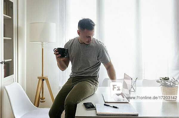 Geschäftsmann hält eine Kaffeetasse und benutzt einen Laptop  während er im Büro zu Hause auf dem Schreibtisch sitzt
