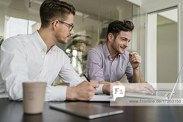 Männlicher Unternehmer  der an einem Laptop arbeitet  während ein Kollege neben ihm im Büro sitzt