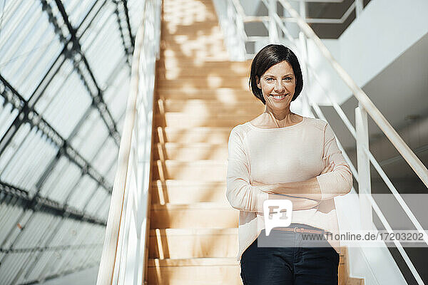 Smiling female professional with arms crossed against steps in corridor