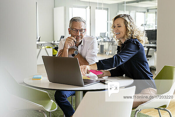 Lächelnde Geschäftsfrau  die auf einen Laptop zeigt  während sie neben einem reifen Geschäftsmann am Tisch sitzt  während einer Besprechung im Büro