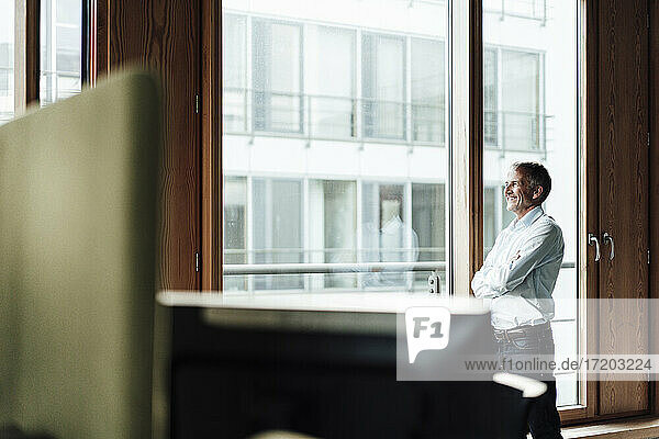 Lächelnder älterer männlicher Unternehmer mit verschränkten Armen  der durch ein Glasfenster im Büro schaut