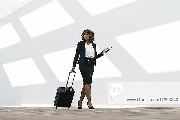 Businesswoman with mobile phone and wheeled luggage walking against white wall