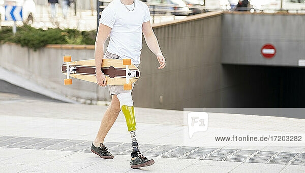 Young disabled man holding skateboard while walking on footpath