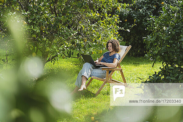 Mittlere erwachsene Frau,  die einen Laptop benutzt und auf einem Stuhl inmitten von Bäumen im Garten sitzt
