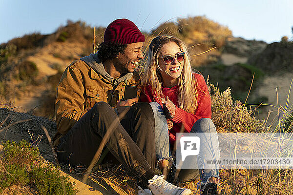 Girlfriend smiling while sitting by cheerful boyfriend on sand dune during sunset