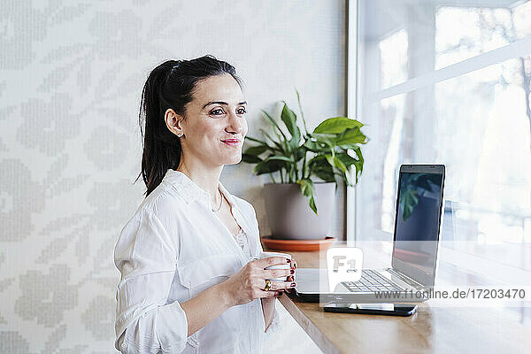 Weibliche Unternehmerin hält eine Tasse Kaffee in der Hand und blickt durch das Fenster eines Cafés
