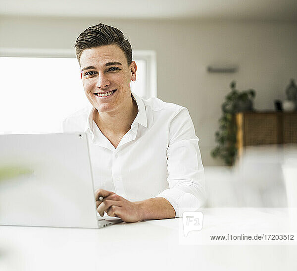 Lächelnde männliche Fachkraft  die einen Laptop benutzt  während sie an einem Tisch im Heimbüro sitzt