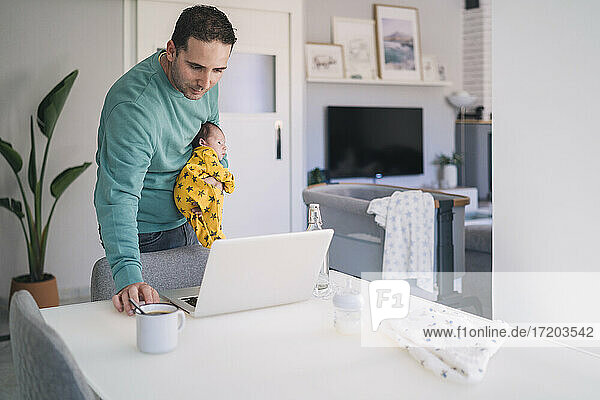Vater trägt seinen Sohn  während er im Büro zu Hause auf den Laptop schaut