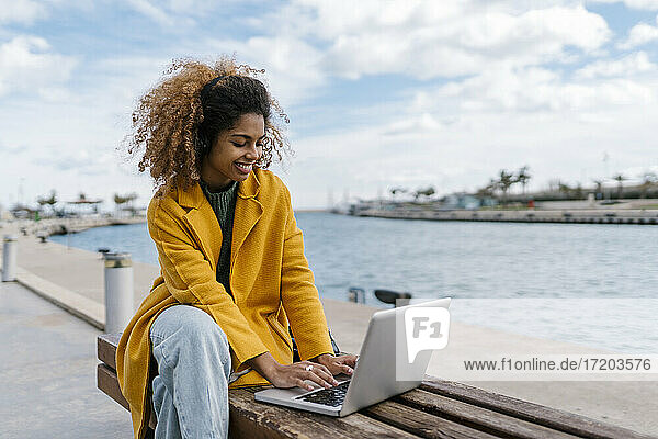 Lächelnde Afro-Frau  die einen Laptop benutzt  während sie auf einer Bank am Fluss sitzt