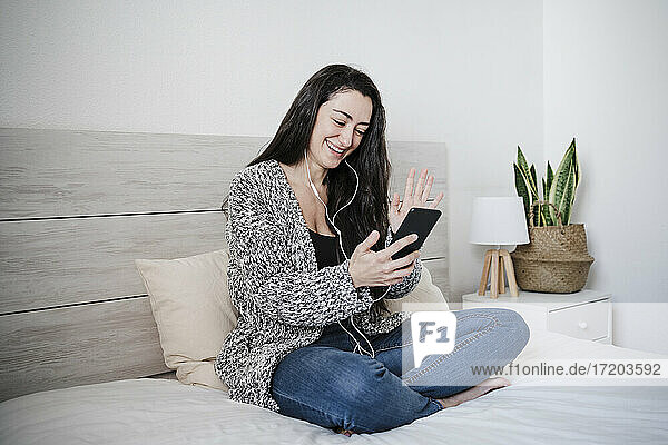 Lächelnde Frau winkt mit der Hand während eines Videoanrufs über ein Mobiltelefon auf einem Bett an der Wand zu Hause