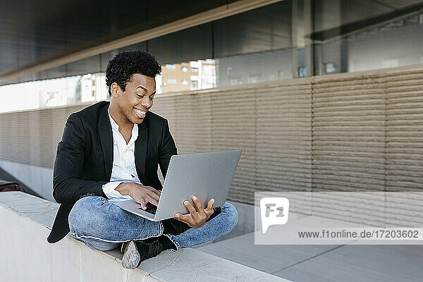 Smiling businessman using laptop while sitting cross legged on retaining wall