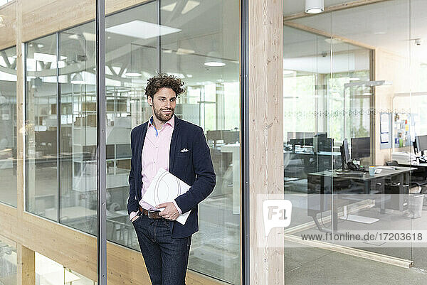 Male entrepreneur holding document while leaning on glass wall in factory