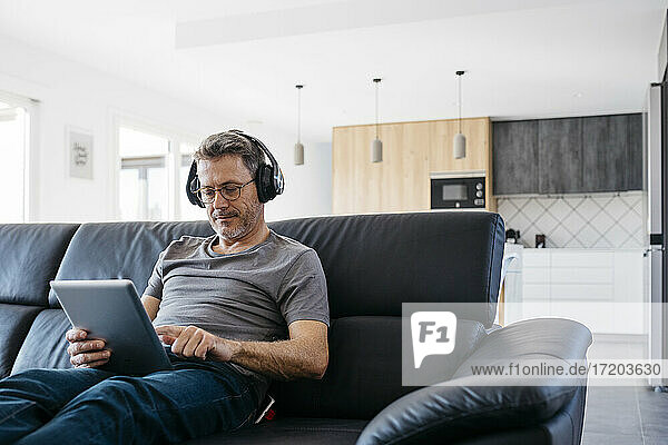 Mature man with headphones using digital tablet while sitting on sofa at home