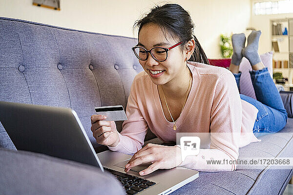 Young woman with credit card smiling while using laptop at home