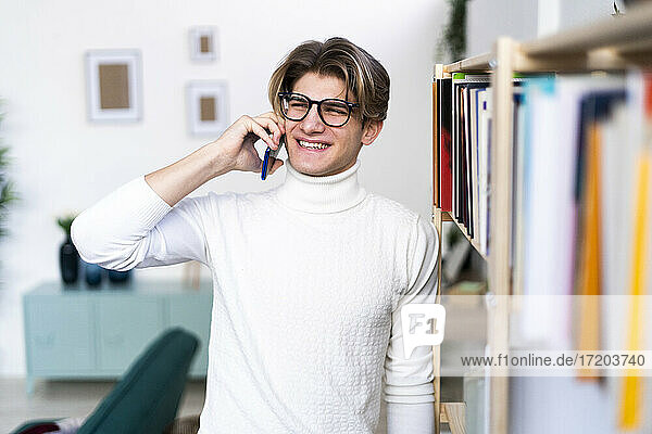 Happy young man talking on phone while standing by bookshelf in living room