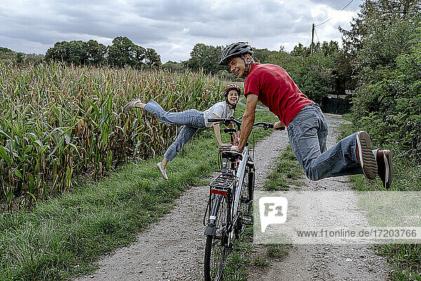 Mature man with daughter jumping while leaning in bicycle at agricultural field