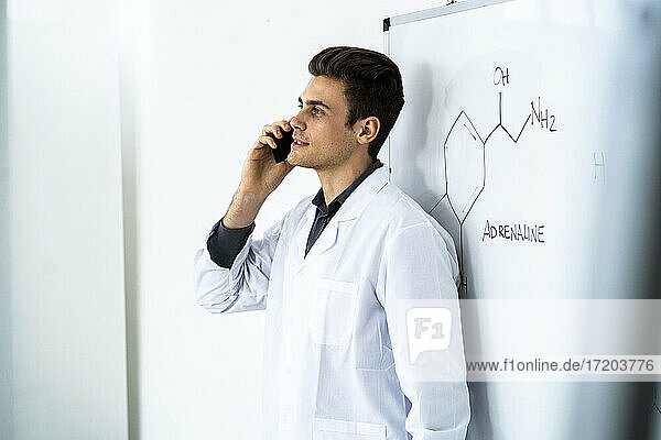 Junger männlicher Wissenschaftler  der mit seinem Smartphone spricht  während er vor einer Tafel im Labor steht