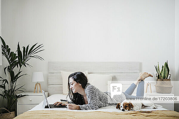 Frau mit Kreditkarte beim Online-Einkauf über den Laptop  während sie mit ihrem Jack Russell Terrior zu Hause auf dem Bett liegt