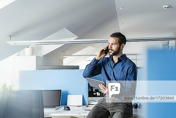 Berufstätiger mit digitalem Tablet  der im Büro sitzend mit seinem Mobiltelefon spricht