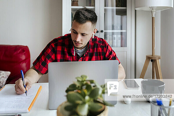 Handsome male entrepreneur with in-ear headphones writing while working on laptop at home office