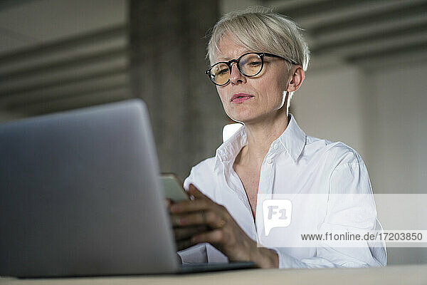 Businesswoman with laptop using smart phone while sitting at home office