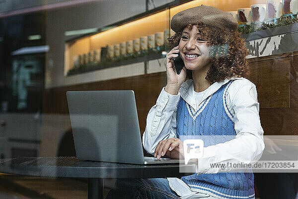 Lächelndes Teenager-Mädchen mit Baskenmütze  das mit einem Laptop in einem Café sitzt und mit einem Mobiltelefon spricht