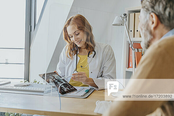 Lächelnder Arzt  der Patienten über Röntgenbilder aufklärt  während er an der Trennwand in der Arztpraxis sitzt