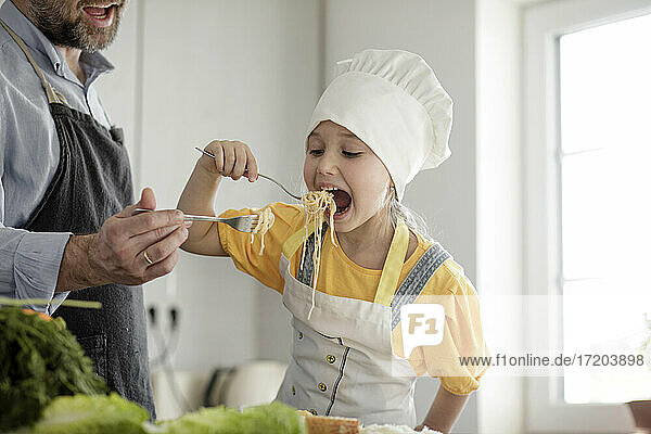 Mädchen mit Schürze und Kochmütze isst Nudeln mit Vater in der Küche zu Hause