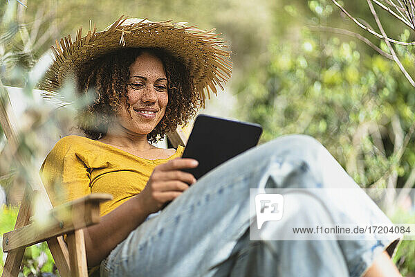 Smiling woman in straw hat using digital tablet while sitting on chair in garden