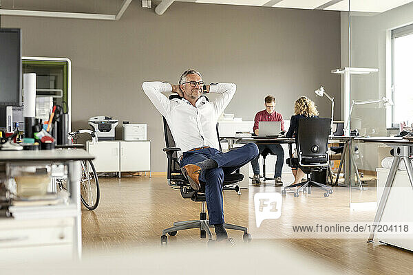 Lächelnder männlicher Geschäftsmann mit Händen hinter dem Kopf  der wegschaut  während er auf einem Stuhl im Büro sitzt