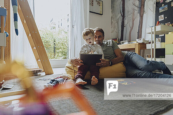 Father lying on bean bag while looking at son using tablet in bedroom