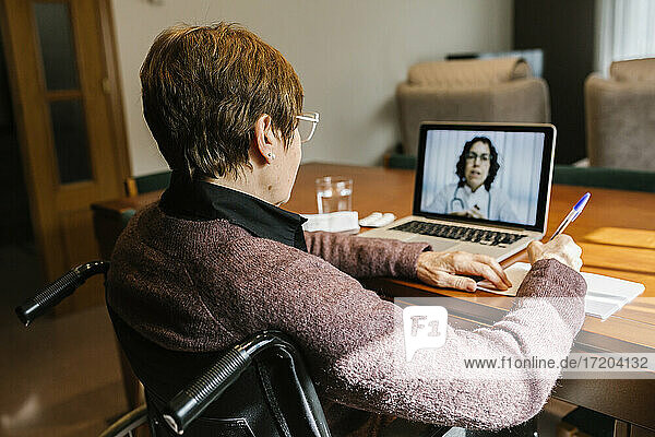 Ältere Frau sitzt im Rollstuhl und schreibt in ein Buch  während sie zu Hause eine Videokonsultation während COVID-19 durchführt