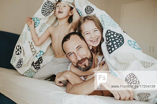Fröhliche Tochter mit Vater  während der Bruder unter der Decke auf dem Bett zu Hause wegschaut