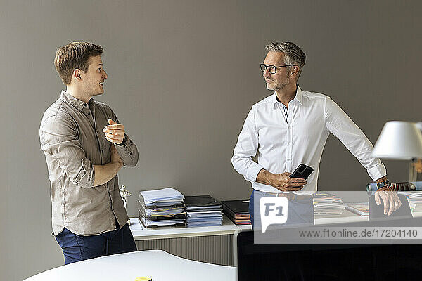 Young businessman talking with mature colleague while standing against wall in office